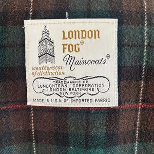 Puede incluir: Primer plano de una etiqueta de prenda London Fog Maincoats. La etiqueta crema muestra el nombre de la marca, el logotipo y la frase "weatherwear of distinction". La etiqueta est&aacute; cosida sobre una tela a cuadros verde, marr&oacute;n y roja. Hecho en EE. UU.