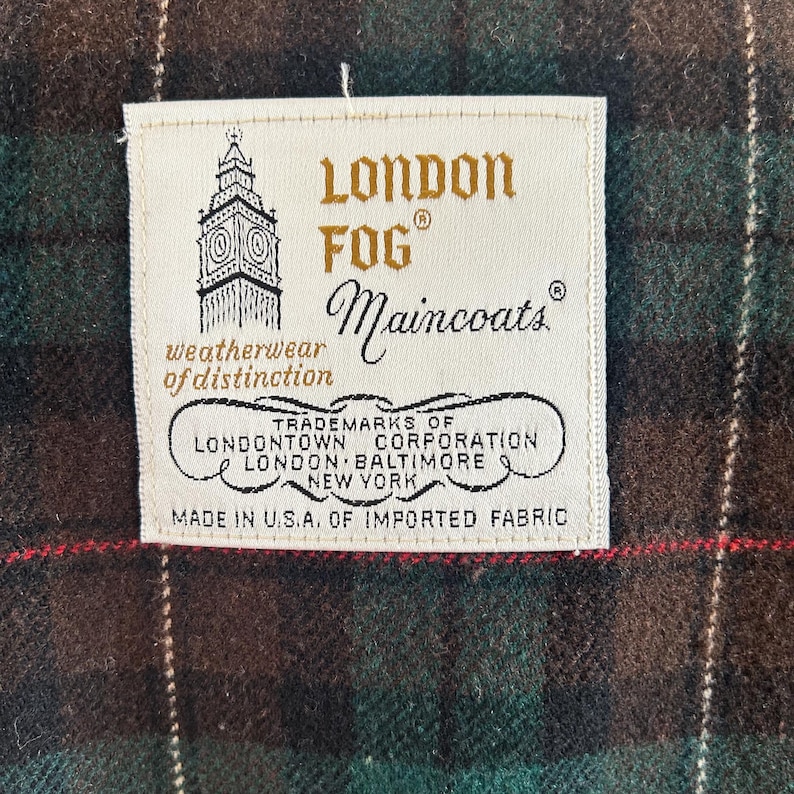 Puede incluir: Primer plano de una etiqueta de prenda London Fog Maincoats. La etiqueta crema muestra el nombre de la marca, el logotipo y la frase "weatherwear of distinction". La etiqueta est&aacute; cosida sobre una tela a cuadros verde, marr&oacute;n y roja. Hecho en EE. UU.