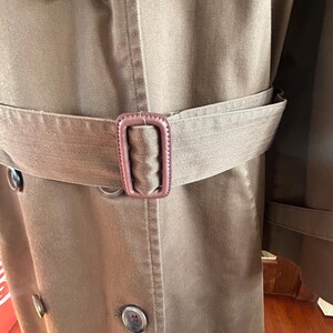 Puede incluir: Primer plano de una gabardina marr&oacute;n con cintur&oacute;n y hebilla a juego. El abrigo presenta un dise&ntilde;o cruzado con botones grandes y un cuello cl&aacute;sico. El cintur&oacute;n se ajusta a la cintura, resaltando la silueta entallada del abrigo.