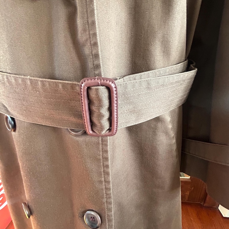 Puede incluir: Primer plano de una gabardina marr&oacute;n con cintur&oacute;n y hebilla a juego. El abrigo presenta un dise&ntilde;o cruzado con botones grandes y un cuello cl&aacute;sico. El cintur&oacute;n se ajusta a la cintura, resaltando la silueta entallada del abrigo.