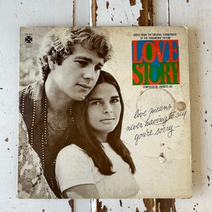 以下が含まれることがあります： パラマウント映画のオリジナルサウンドトラックからの音楽、ラブストーリーのヴィンテージビニールレコードカバー。モノクロ写真で、抱き合っているカップルが写っている。カバーには「ラブストーリー」というタイトルと、「Music from the original soundtrack of the Paramount Picture」と「Composed by Francis Lai」というテキストが書かれている。タイトルの下には、「love means never having to say you're sorry」というテキストが筆記体で書かれている。