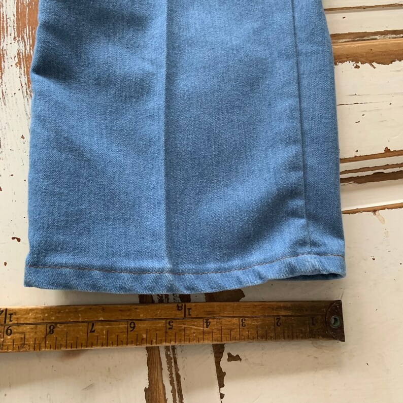 May include: Close-up of the bottom hem of a pair of blue denim pants. The hem is sewn with a straight stitch and the fabric is a light blue wash.