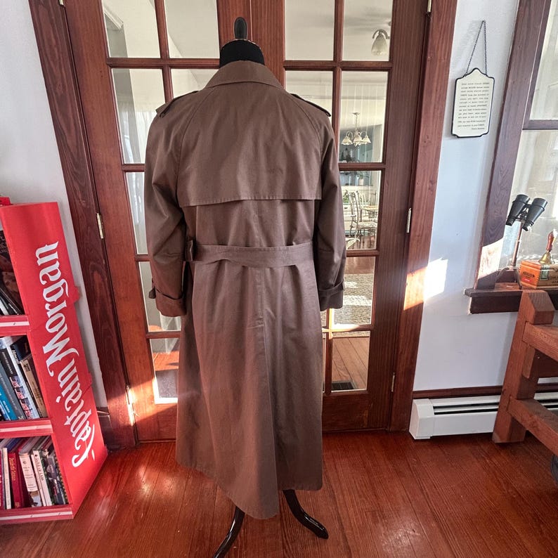 Puede incluir: Un trench coat marr&oacute;n exhibido en un maniqu&iacute;. El abrigo presenta un dise&ntilde;o cruzado, un cintur&oacute;n en la cintura y charreteras en los hombros. La tela es suave y duradera. Una prenda de abrigo cl&aacute;sica.