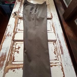 Puede incluir: Un pantalón de vestir marrón, colocado sobre una superficie de madera blanca desgastada. El pantalón tiene un diseño clásico con cierre de botón y cremallera, y bolsillos. La tela parece ser un material de peso medio, adecuado para diversas ocasiones.
