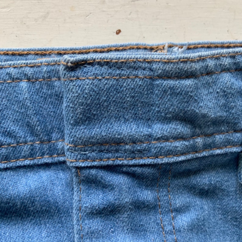 May include: Close-up of the waistband of a pair of blue denim jeans. The waistband is folded over and has a double row of stitching.