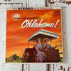 Puede incluir: Portada de álbum vintage de Capitol Records para "Oklahoma!" de Rodgers y Hammerstein, con una pareja en un carruaje tirado por caballos contra un cielo naranja. La portada incluye el texto "CAPITOL STEREO" y "THE FULL SPECTRUM OF SOUND".