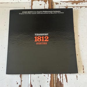 May include: A black vinyl record album cover with the title "Tchaikovsky 1812 Overture" in red text. The album features the Los Angeles Philharmonic Orchestra conducted by Zubin Mehta. The album cover also includes the text "Original Civil War cannon, bells and carillon by Maas-Row Carillons."
