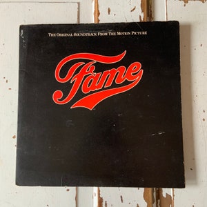Puede incluir: Carátula de disco de vinilo negro con el título "The Original Soundtrack From The Motion Picture Fame" en texto blanco y la palabra "Fame" en texto rojo en una fuente estilizada.