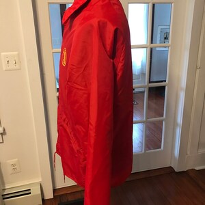 May include: A red windbreaker jacket with a yellow logo on the left chest. The jacket has a zipper closure and a drawstring hood.