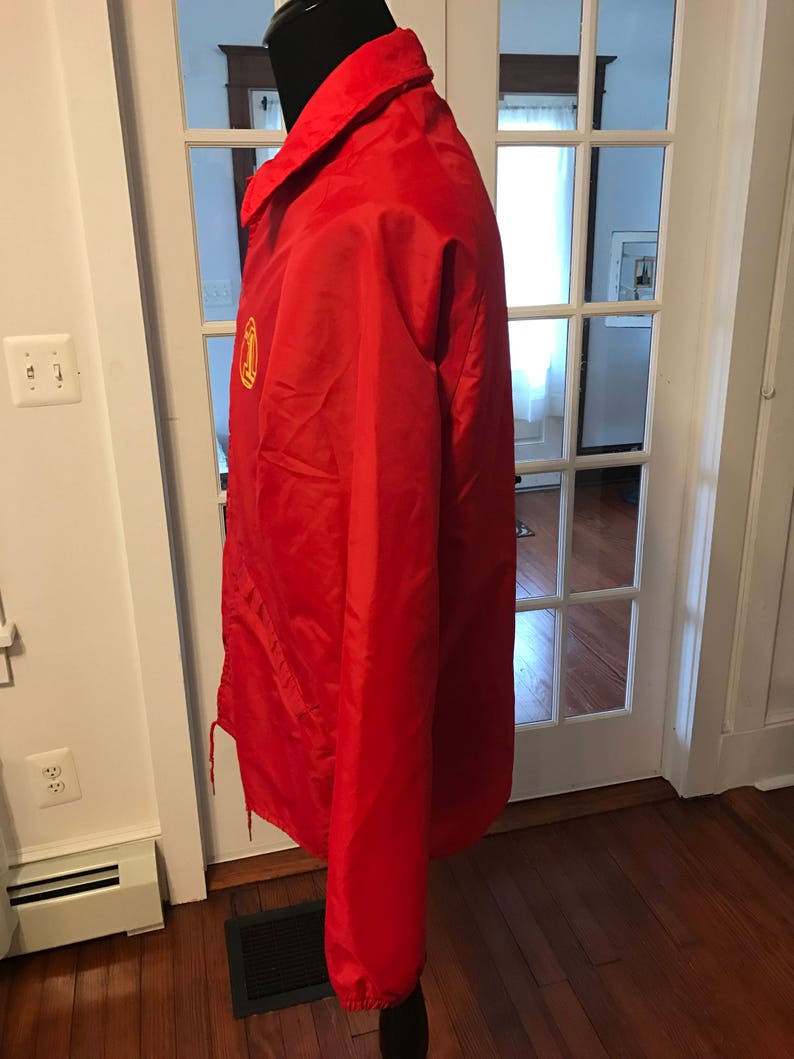 May include: A red windbreaker jacket with a yellow logo on the left chest. The jacket has a zipper closure and a drawstring hood.
