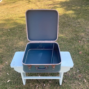 Puede incluir: Una maleta vintage azul con un asa de metal y un cierre. La maleta está abierta y vacía, revelando un interior azul. La maleta está sentada en un banco de madera blanco.