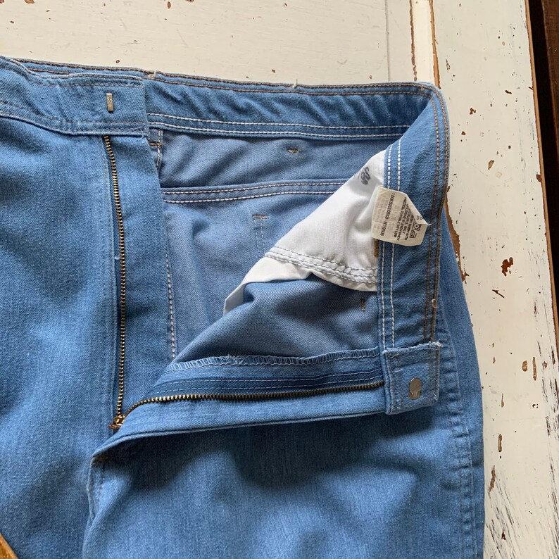 May include: A pair of light blue denim jeans with a zipper fly and a button closure. The jeans have a white label sewn into the waistband with the text "Made in Bangladesh" and "100% Cotton".