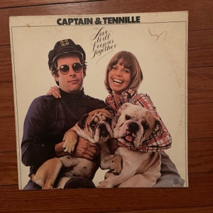 Puede incluir: Una portada de álbum de vinilo vintage con Captain & Tennille. La portada presenta una foto del dúo con dos bulldogs. El título del álbum es "Love Will Keep Us Together".