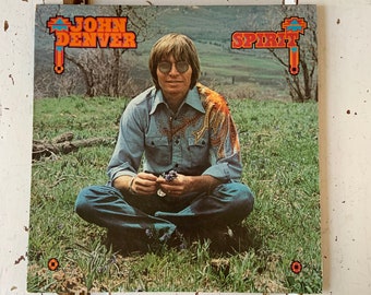 May include: A vintage vinyl record album cover featuring John Denver. The cover art shows John Denver sitting in a field of green grass, holding a small bouquet of purple flowers. The album title "Spirit" is displayed in orange letters on the top right corner of the cover.