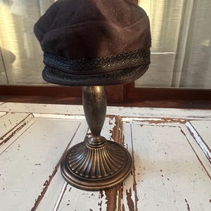 May include: A dark brown cloche hat with a decorative black trim, displayed on a bronze-coloured stand. The hat has a rounded crown and a structured brim. The stand has a detailed, ornate base.