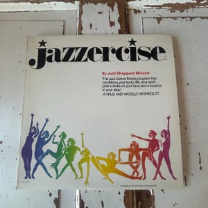 May include: A vintage Jazzercise record album cover featuring a group of women in silhouette poses, wearing colorful outfits, against a white background. The album title "Jazzercise" is in bold black letters with a star on either side. The text "By Judi Sheppard Missett" is below the title, followed by a description of the fitness program. The text "Includes a 20x40 instructional poster" is at the bottom of the cover.