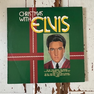 Peut inclure: Un album vinyle vintage intitulé "Christmas With Elvis". La couverture de l'album est verte avec des rubans rouges et le texte "Christmas With Elvis" en jaune. Un portrait d'Elvis Presley est au centre. Les titres des chansons sont listés en bas.