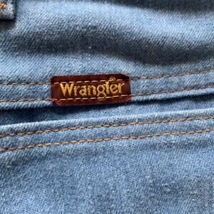 May include: Close-up of a blue denim fabric with a brown leather patch that says "Wrangler" in gold lettering.