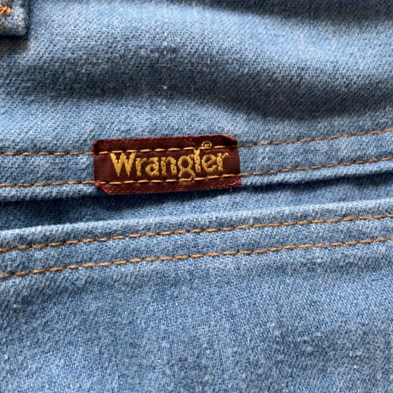 May include: Close-up of a blue denim fabric with a brown leather patch that says "Wrangler" in gold lettering.