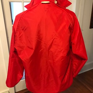 May include: A red nylon jacket with a zipper closure and a stand-up collar. The jacket has long sleeves and a single pocket on the left chest.