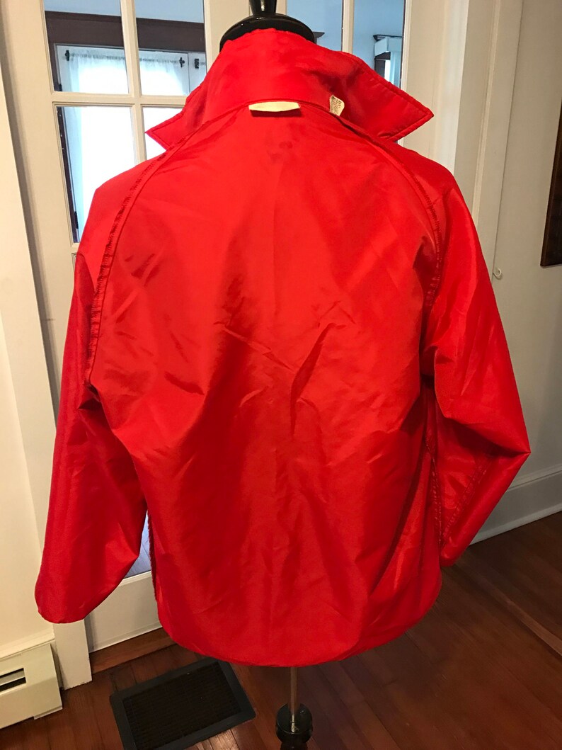 May include: A red nylon jacket with a zipper closure and a stand-up collar. The jacket has long sleeves and a single pocket on the left chest.