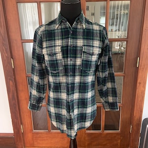 Puede incluir: Una camisa de manga larga a cuadros con un patrón verde, blanco y negro. La camisa tiene cuello abotonado y dos bolsillos en el pecho.