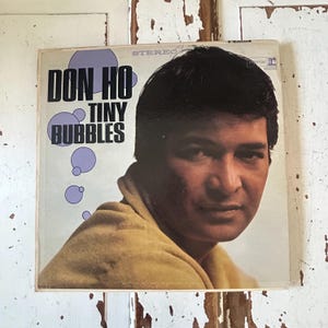 May include: Vintage vinyl record album cover featuring Don Ho and the title "Tiny Bubbles." The cover has a light background with the album title in bold black letters and purple bubble graphics. The image shows a man in a yellow shirt.