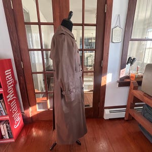 Puede incluir: Un abrigo tipo trench marr&oacute;n expuesto en un maniqu&iacute;. El abrigo presenta un dise&ntilde;o cruzado, un cintur&oacute;n en la cintura y una longitud larga. El abrigo est&aacute; hecho de una tela suave y tiene un estilo cl&aacute;sico y atemporal. El abrigo se exhibe frente a una puerta de madera.