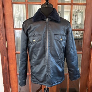 Può includere: Una giacca di raso blu scuro con chiusura a zip e colletto in pelliccia sintetica. La giacca ha due tasche a patta con bottoni dorati.