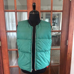 May include: A green puffer vest with a full-length silver zipper. The vest has a black collar and red lining visible at the bottom. The vest is displayed on a black mannequin.