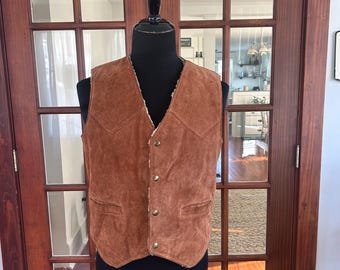 May include: A brown suede vest with a cream-colored lining and gold-colored buttons. The vest has two front pockets and a V-neck design. The vest is displayed on a black mannequin.