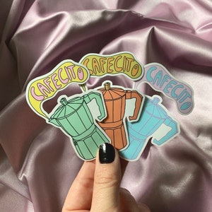 May include: Three colourful stickers featuring a cartoon illustration of a coffee pot with the word "Cafecito" written in a whimsical font.