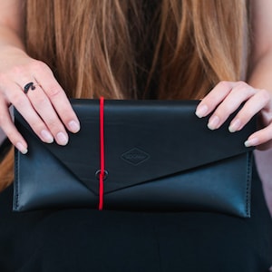 Könnte beinhalten: Eine schwarze Leder-Kupplung in Briefform mit einem roten Gummibandverschluss und einem kleinen rautenförmigen Logo mit der Aufschrift "SOOMVA".