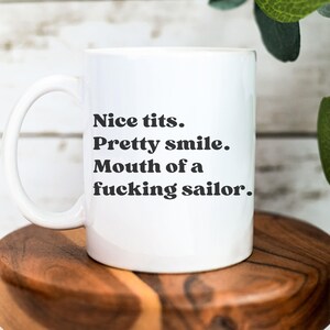 May include: White ceramic mug with a curved handle. The mug features black text that reads: "Nice tits. Pretty smile. Mouth of a f***ing sailor."