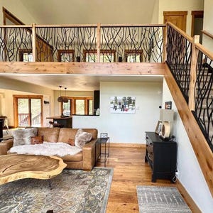 May include: Interior view of a living room with a loft and staircase. The railing features a black, organic branch design. A brown leather sofa, wooden coffee table, and hardwood floors complete the rustic aesthetic. The room is well-lit with natural light.