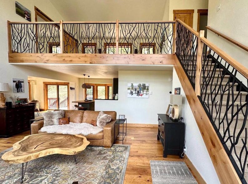 May include: Interior view of a living room with a loft and staircase. The railing features a black, organic branch design. A brown leather sofa, wooden coffee table, and hardwood floors complete the rustic aesthetic. The room is well-lit with natural light.
