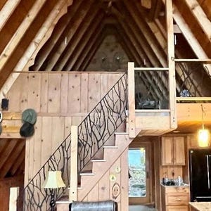 May include: Interior shot of a cabin with a wooden staircase featuring a decorative black metal railing. The cabin has a high, sloped ceiling with exposed wooden beams. A black leather armchair sits near the stairs, and a lamp provides soft lighting.