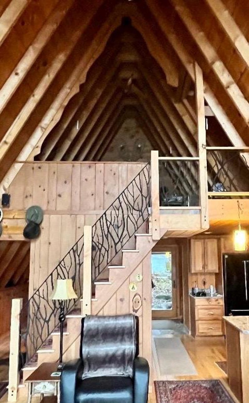 May include: Interior shot of a cabin with a wooden staircase featuring a decorative black metal railing. The cabin has a high, sloped ceiling with exposed wooden beams. A black leather armchair sits near the stairs, and a lamp provides soft lighting.