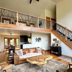 May include: A spacious living room with a loft and staircase. The railing features a black, tree-branch design. A brown leather sofa, wooden coffee table, and patterned rug complete the cozy, rustic interior. The room has a high ceiling and natural light.