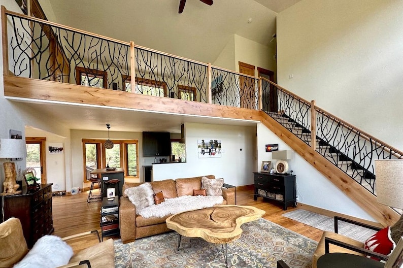 May include: A spacious living room with a loft and staircase. The railing features a black, tree-branch design. A brown leather sofa, wooden coffee table, and patterned rug complete the cozy, rustic interior. The room has a high ceiling and natural light.