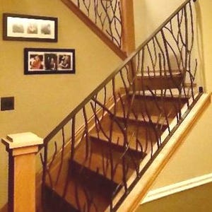 May include: A wooden staircase with a decorative wrought iron railing. The railing has a branch-like design. The stairs are made of wood and lead upwards. The walls are painted a warm beige color. Two framed pictures hang on the wall.
