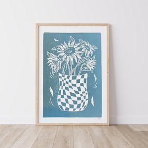 May include: A blue and white linocut print of a vase with a chequered pattern filled with sunflowers. The print is framed in a natural wood frame.