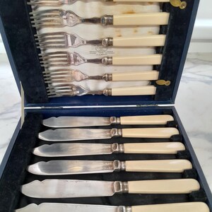 May include: A vintage cutlery set presented in a blue velvet-lined box. The set comprises six forks and six knives. The forks and knives feature cream-coloured handles and silver blades and tines. The box is open, displaying the arrangement of the cutlery.