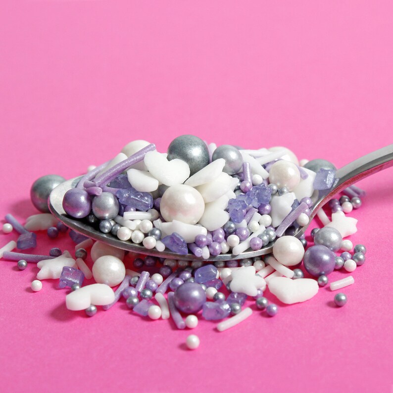Purple, silver and white cake sprinkles featuring edible stars, hearts, sugar strands, nonpareils, sugar rocks and pearls on a spoon.