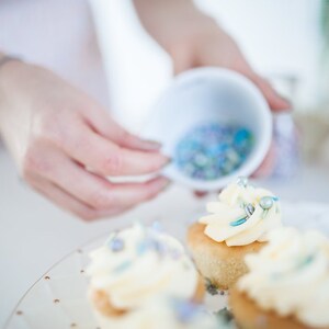 Person decorating 4 cupcakes with butter cream and edible sprinkles.