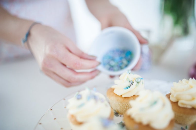 Person decorating 4 cupcakes with butter cream and edible sprinkles.