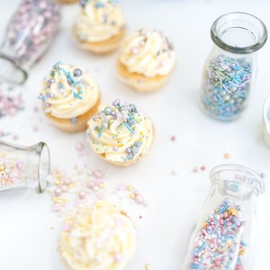 5 cupcakes topped with buttercream and edible cake sprinkles. Also 4 glass jars and one white dish containing cake sprinkles.