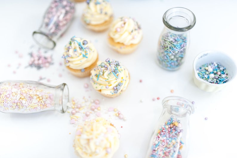 5 cupcakes topped with buttercream and edible cake sprinkles. Also 4 glass jars and one white dish containing cake sprinkles.