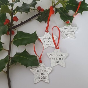 May include: Four silver star-shaped Christmas ornaments with red ribbon. The ornaments have the following text: "GINgle all the way", "gin the Christmas spirit", "let the fun beGIN", and "Oh come let us adore gin".