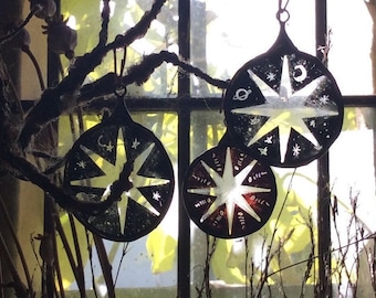 May include: Three glass ornaments with a star design and white paint. The ornaments are hanging from a branch in front of a window.